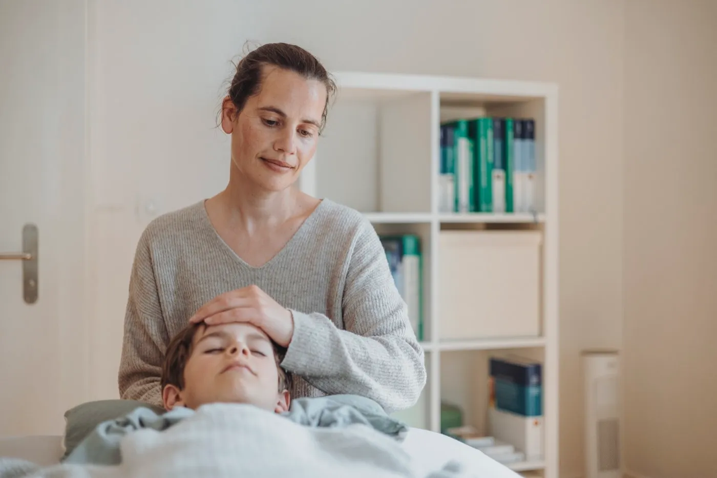 Therapeutin legt eine Hand auf die Stirn eines Kindes, das ruhig auf der Behandlungsliege liegt