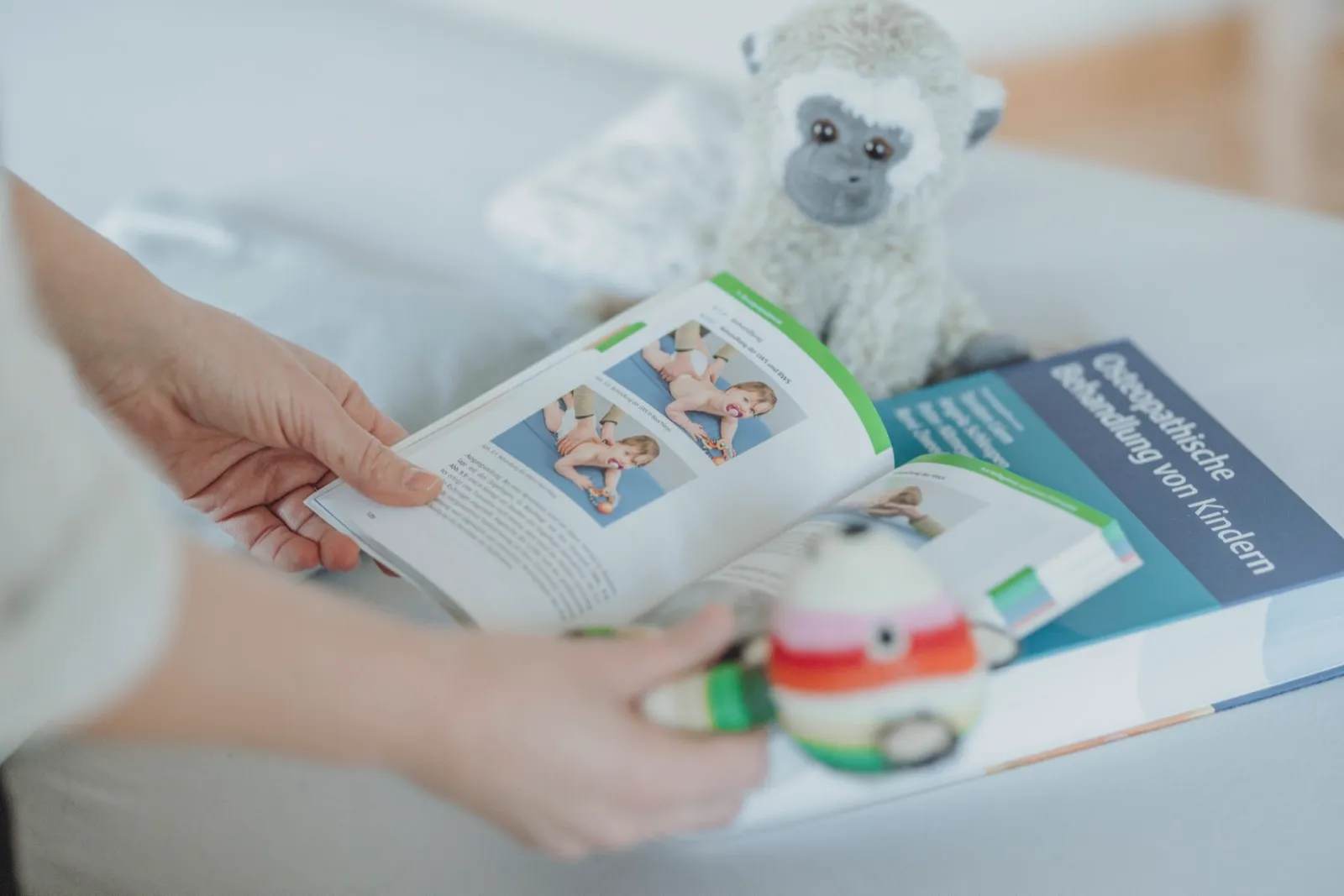 Hände blättern in einem Fachbuch zur osteopathischen Behandlung von Kindern, daneben Kuscheltier und Rassel