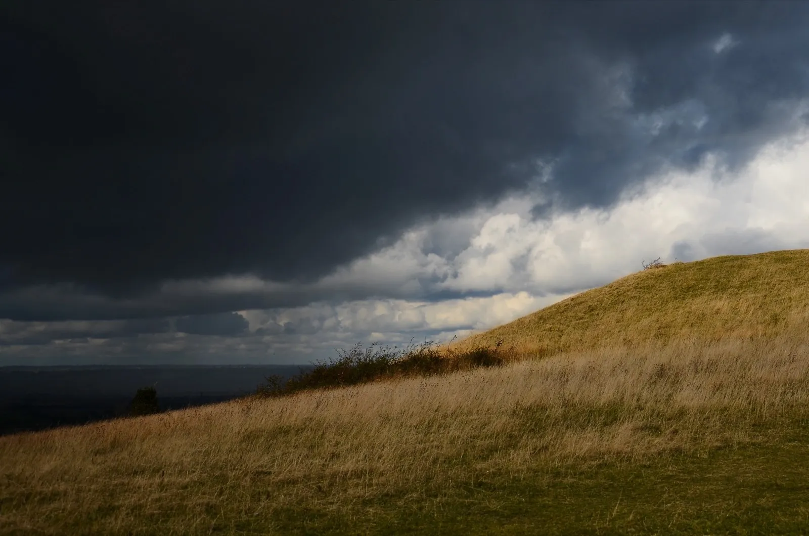 Grasbewachsener Hügel unter dunklen Wolken, am Horizont bricht Licht durch