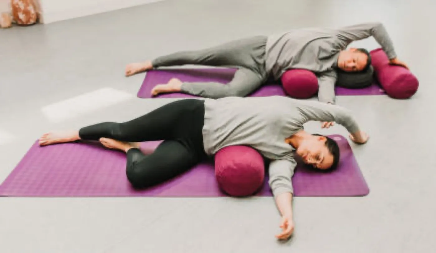 Zwei Personen liegen seitlich über einem Bolster, der obere Arm ist weit über die Flanke gestreckt