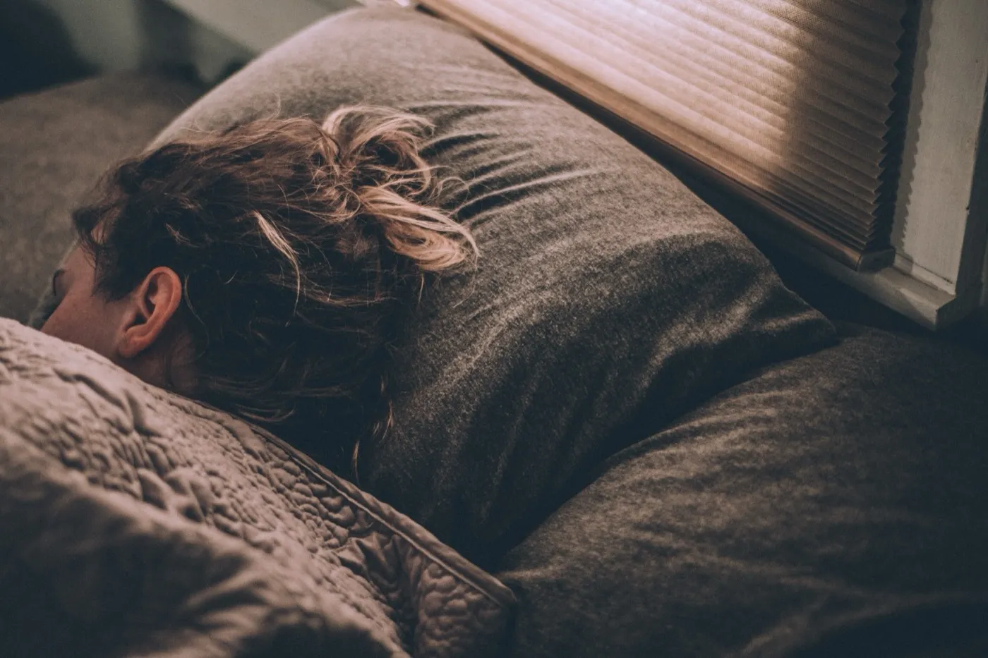 Person schläft eingekuschelt im Bett, gedämpftes Licht fällt durch die Jalousie