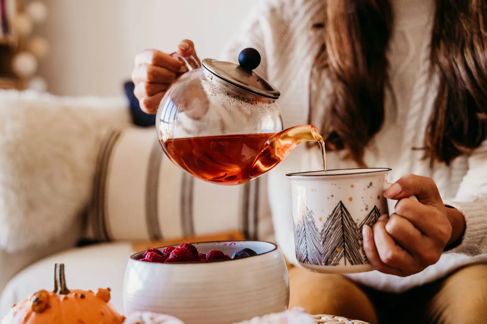 Frau giesst Tee aus einer Glaskanne in eine Tasse, daneben eine Schale mit Beeren
