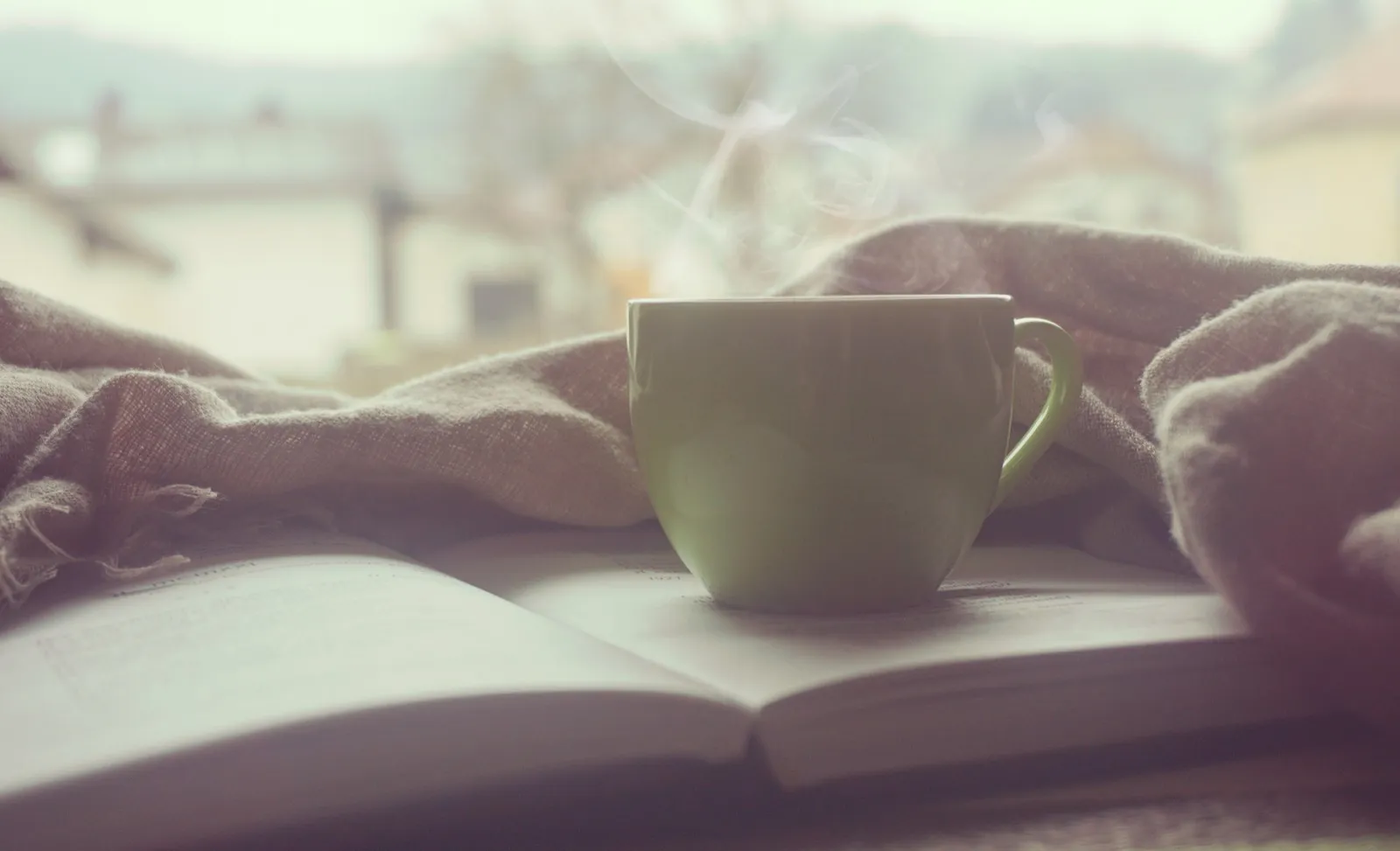 Dampfende Tasse auf einem aufgeschlagenen Buch am Fenster, daneben eine Wolldecke