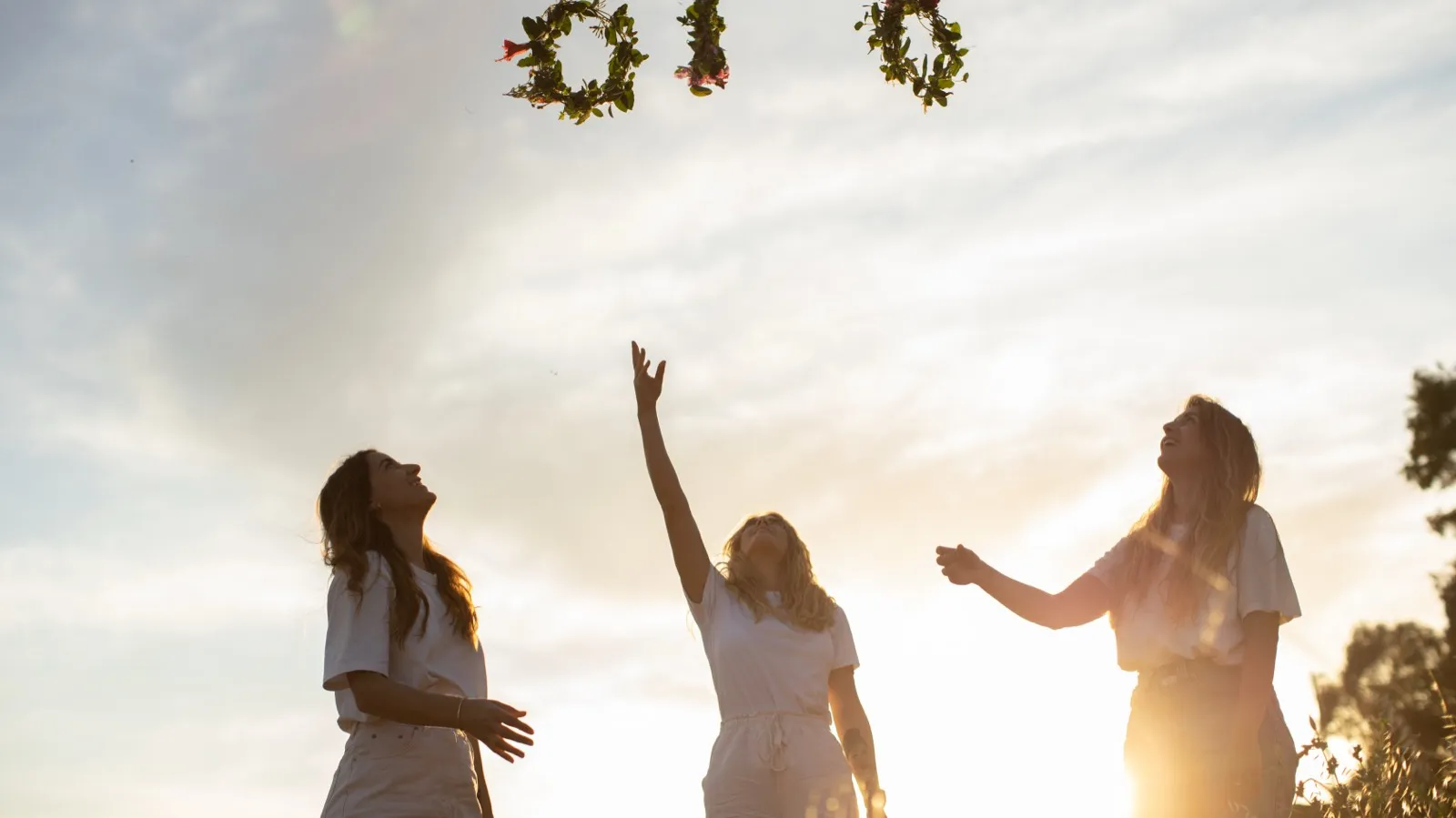 Drei Frauen werfen im Gegenlicht Blumenkränze in die Luft