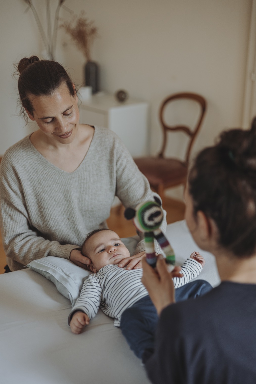 Baby liegt auf der Behandlungsliege und schaut zu einem Spielzeug, die Mutter sitzt lächelnd daneben.