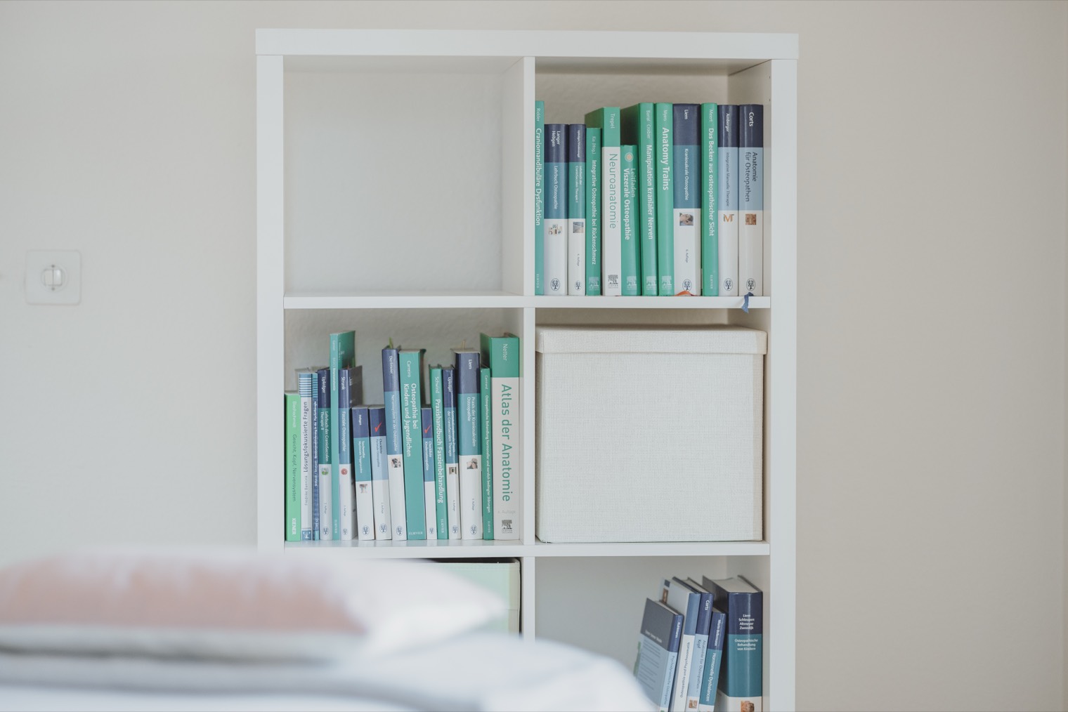 Regal mit Anatomie und Fachbüchern in der Praxis, im Vordergrund eine Behandlungsliege.