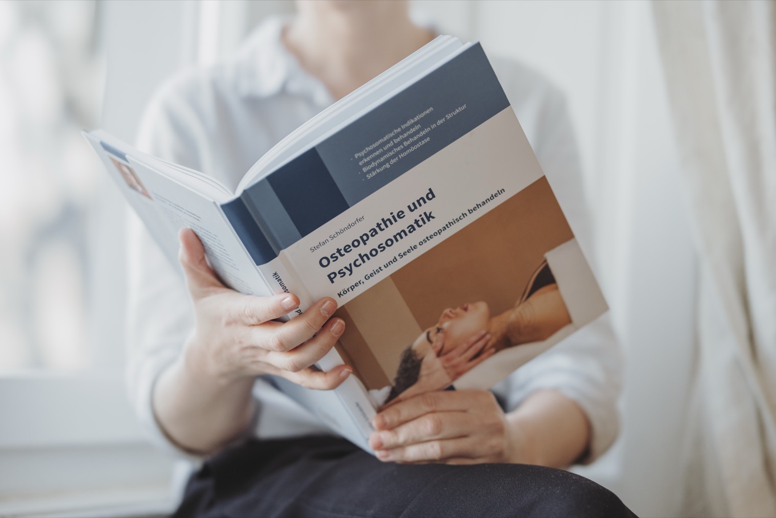 Fachbuch «Osteopathie und Psychosomatik» in den Händen einer Person am Fenster.
